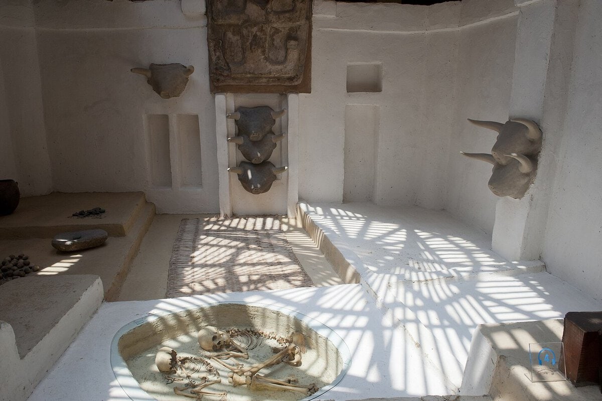Çatalhöyük evinin canlandırmasında taban altına gömülmüş iskelet, arkada boğa başlı duvar
