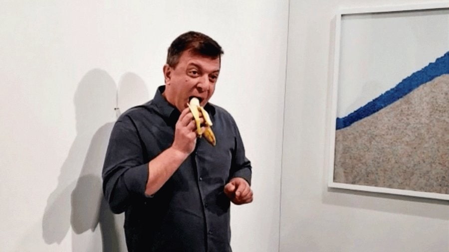 Koyu gömlekli bir adam galeri duvarının önünde soyulmuş bir muzu ısırıyor