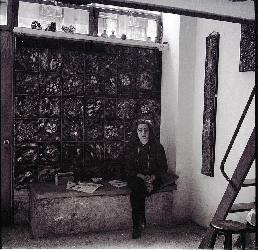 Füreya Koral atölyesinde, arkasındaki koyu sırlı seramik karo panonun önünde taş sedire oturmuş, siyah beyaz fotoğraf
