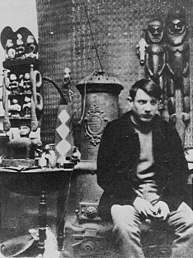 Genç Picasso Montmartre'daki atölyesinde sobanın yanında oturuyor, arkasında koleksiyonundaki Afrika heykelleri