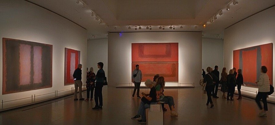 Loş bir sergi salonunda kırmızı ve kahverengi tonlu büyük Rothko tuvallerine bakan ziyaretçiler
