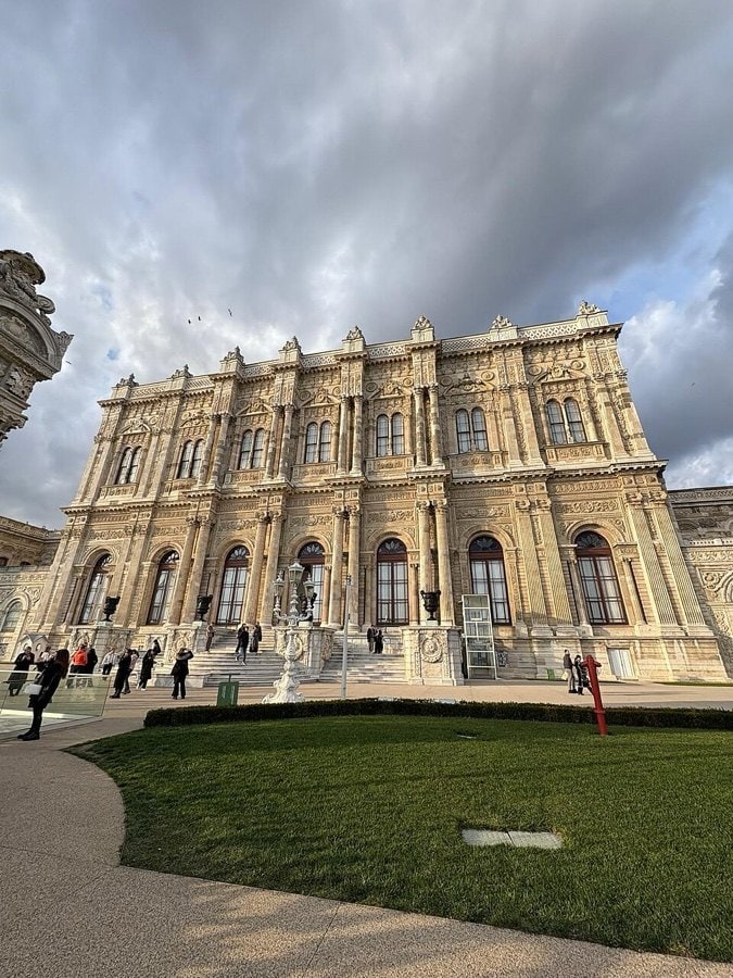 Dolmabahçe Sarayı Selamlık bölümünün süslemeli taş cephesi ve önündeki çimenlik, bulutlu gökyüzü