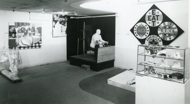 Siyah beyaz sergi fotoğrafı: duvarda Robert Indiana'nın EAT/JUKE/JACK yazılı elmas tablosu, cam vitrinde pasta heykelleri, oturan alçı figür