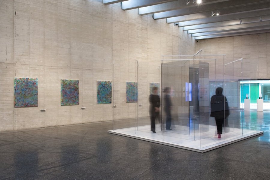Beton duvarlı galeride ziyaretçilerin içinde dolaştığı şeffaf cam labirent, duvarda renkli resimler