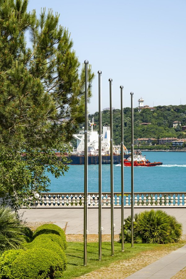 Boğaz kıyısındaki müze bahçesinde yan yana dikilmiş bayraksız metal bayrak direkleri, arkada geçen bir gemi
