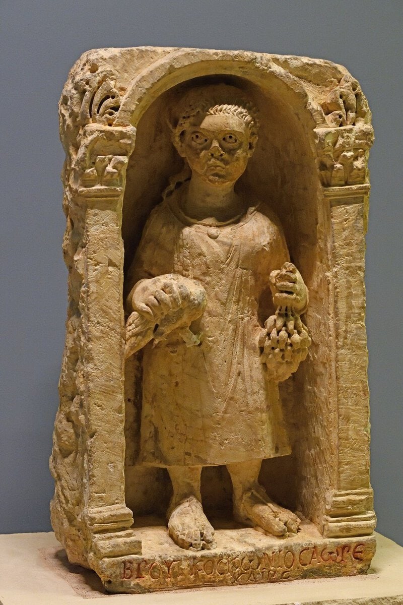 Zeugma Mozaik Müzesi'nde kemerli nişte ayakta duran, bir elinde kuş, diğerinde üzüm salkımı tutan çocuk figürlü Roma mezar steli